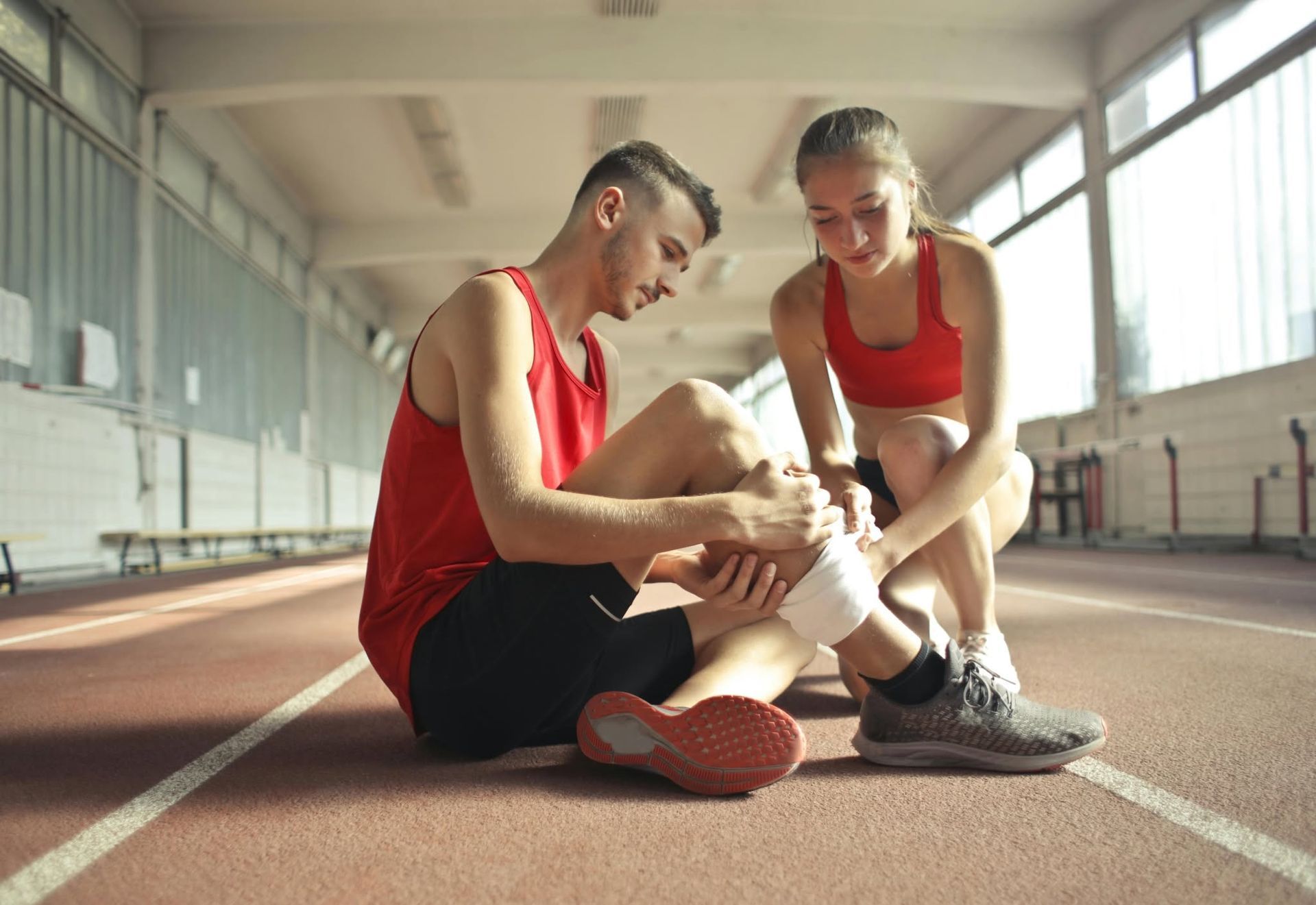 Two runners in red jerseys on an indoor track, one sitting and holding an ankle while the other checks it