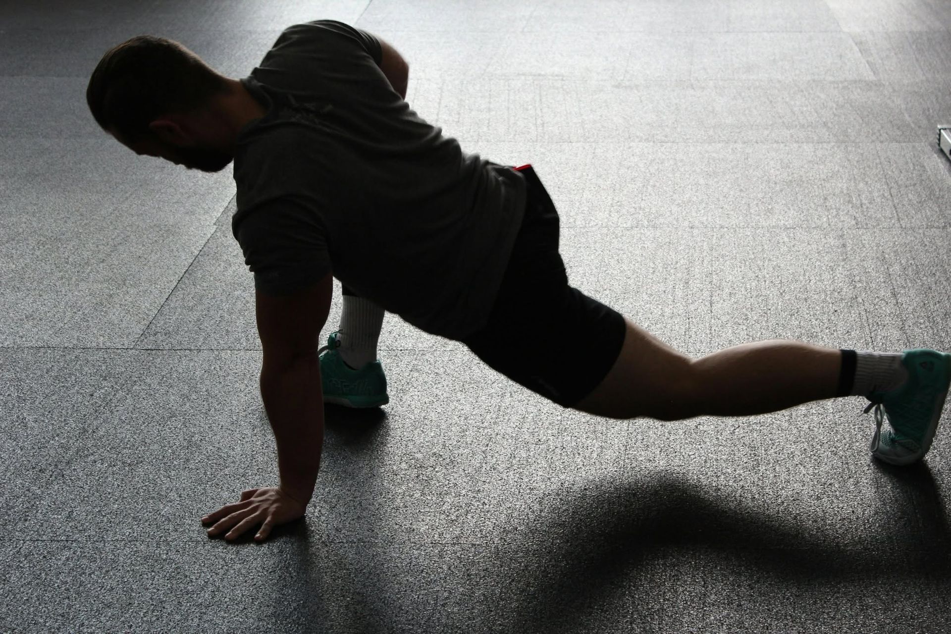 Person stretching in a low lunge on a gym floor, wearing black workout clothes and teal shoes.