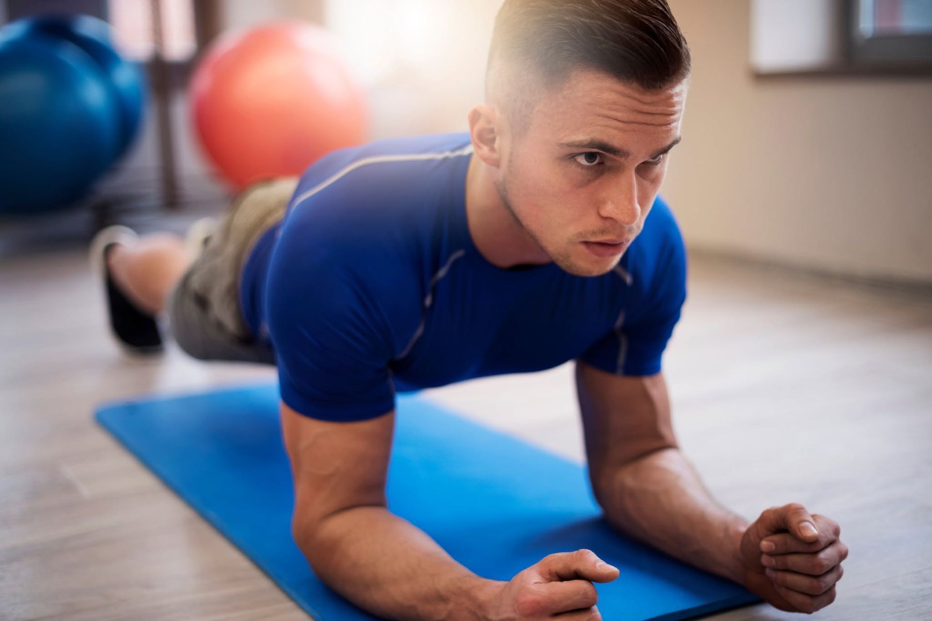 Man doing a forearm plank on a blue mat in a gym, with exercise balls in the background