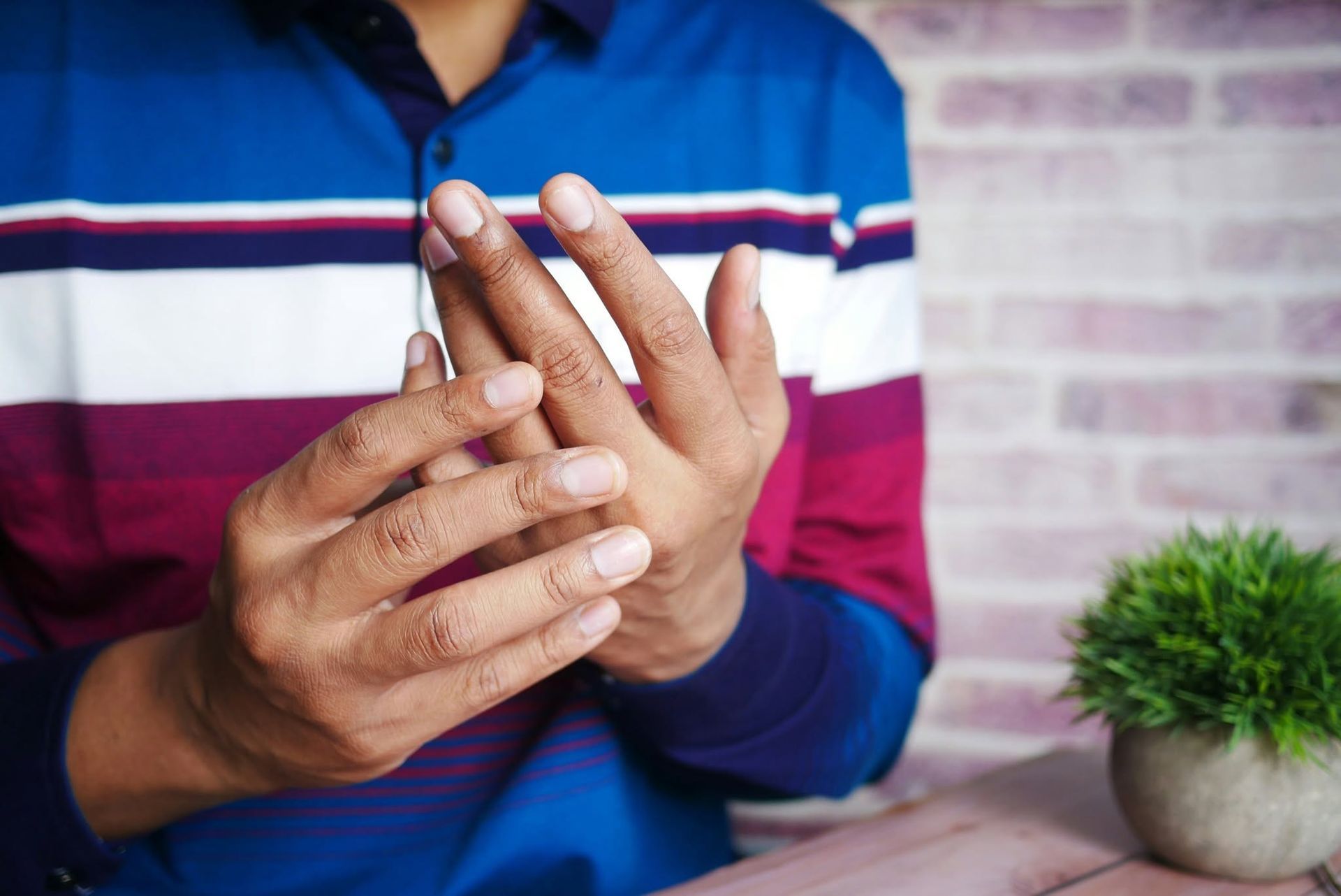 Close-up of hands clasping a finger, with a striped shirt and small potted plant on a table.