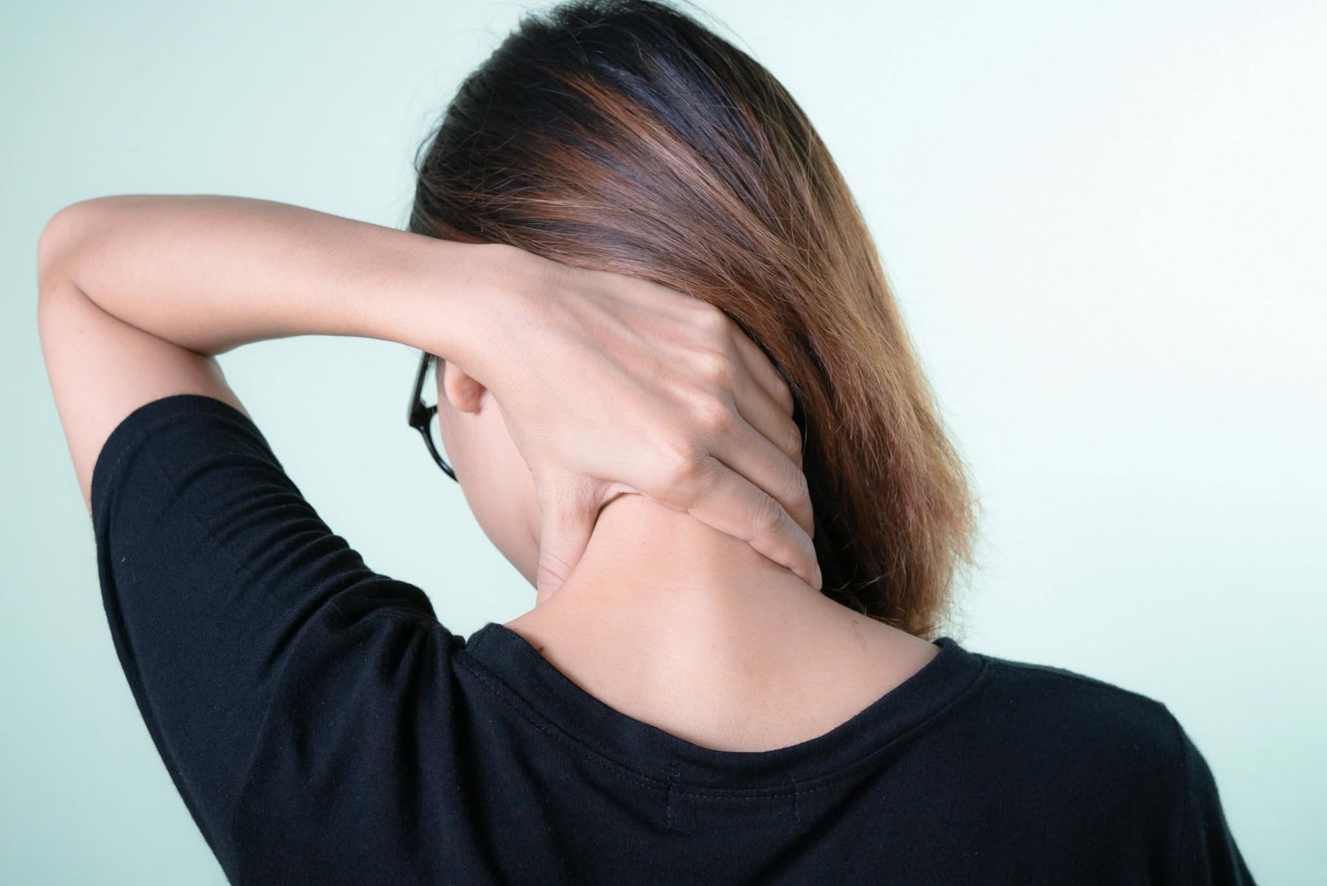 Person holds neck with hand, seen from behind, wearing a black shirt against a light background