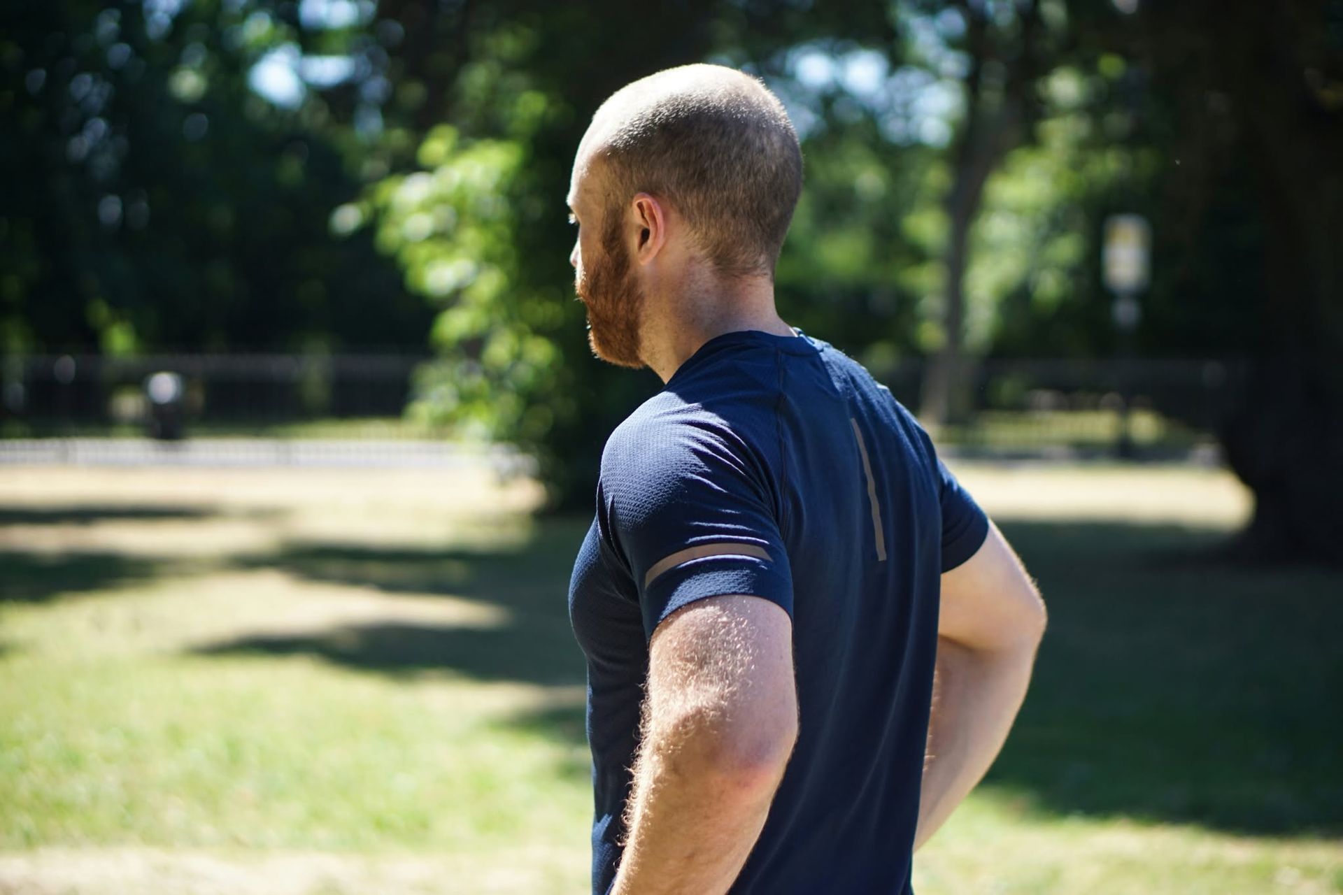 Man in a navy shirt standing in a sunny park, seen from behind with one hand on his hip