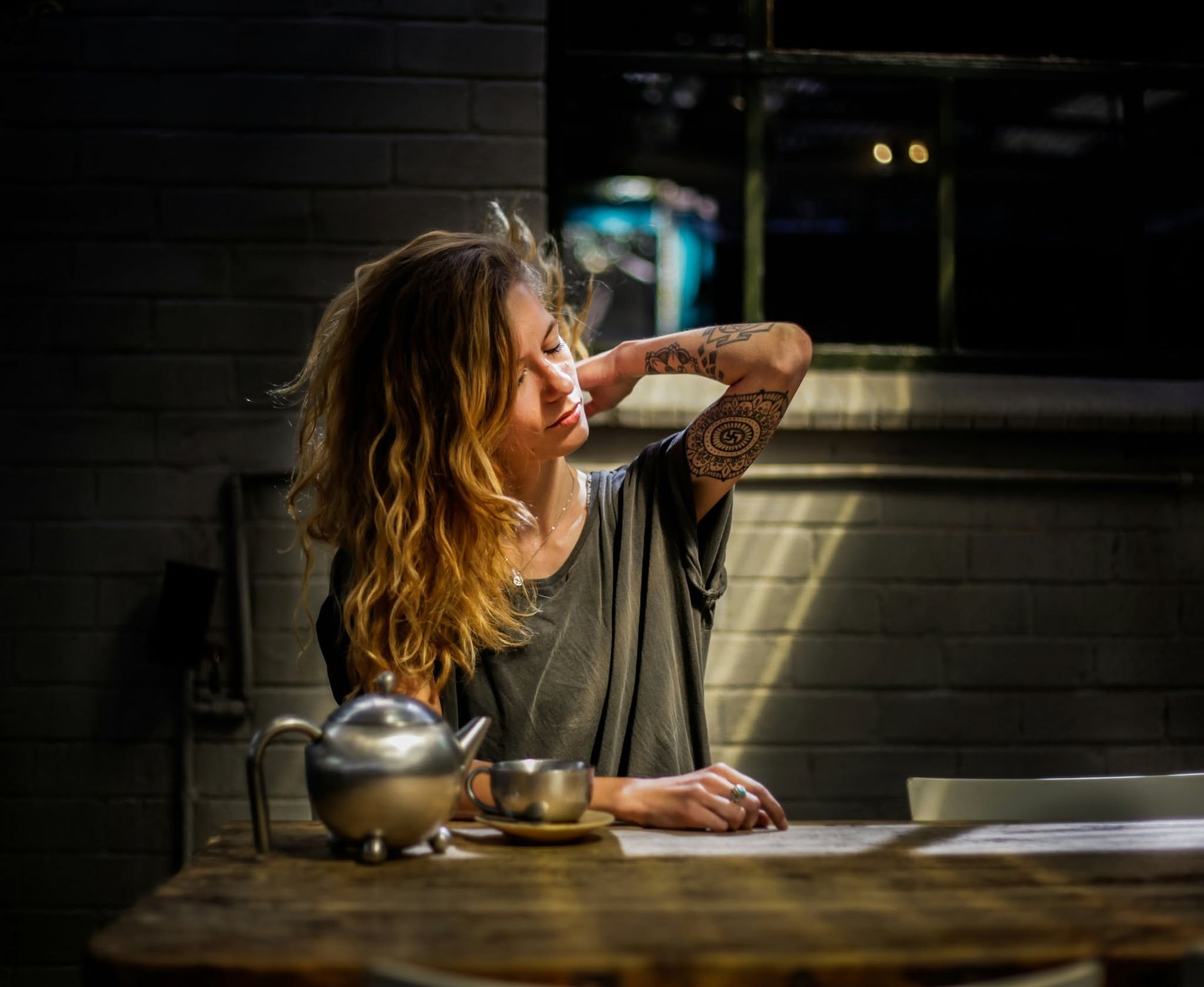 Woman with tattooed arm sitting at a wooden table in a dim cafe, hand in hair, with a teapot and cup.