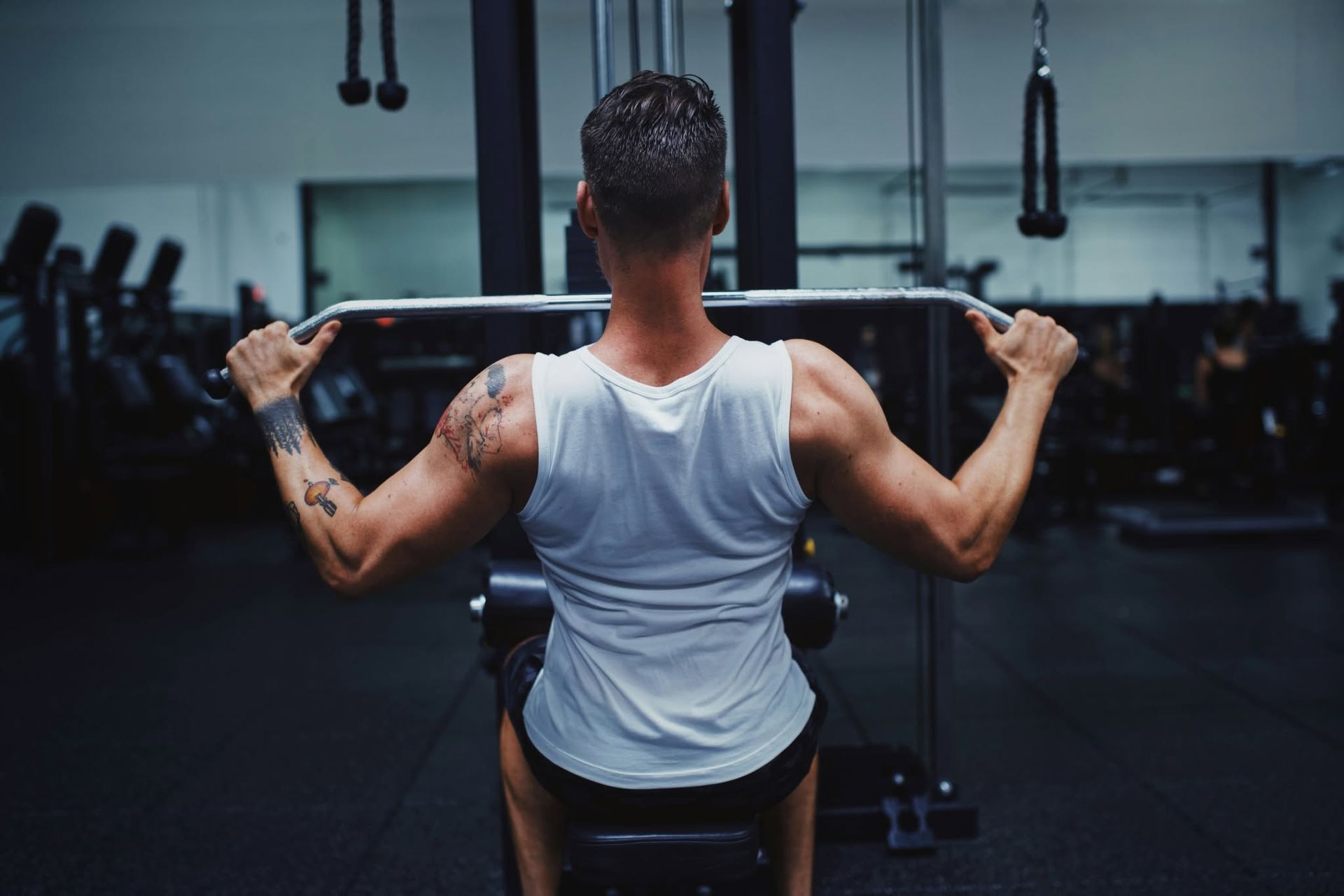 Person lifting a barbell in a gym, seen from behind in a white tank top.