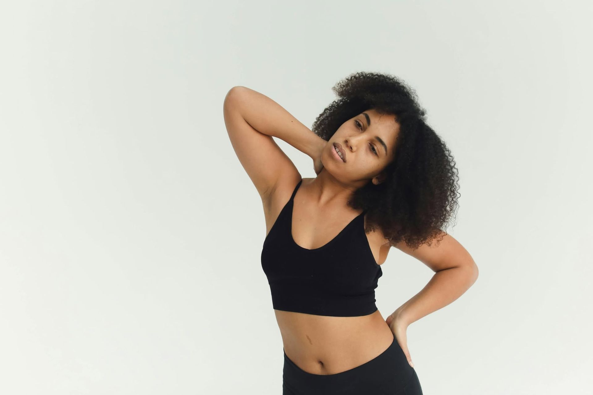 Woman in black sportswear posing against a white background, hand behind head and hip tilted.