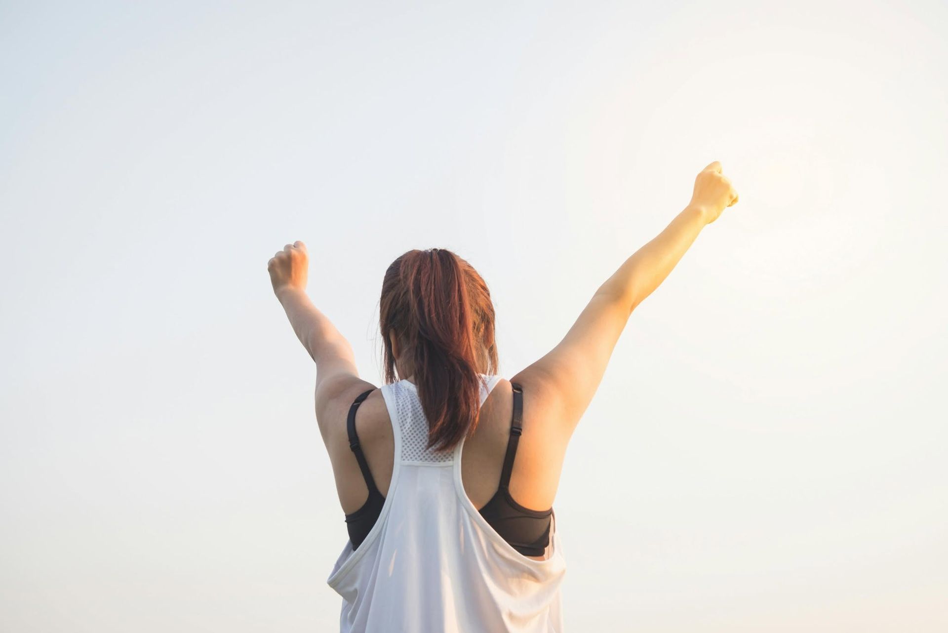 Person in a white tank top raises both arms against a bright sky.