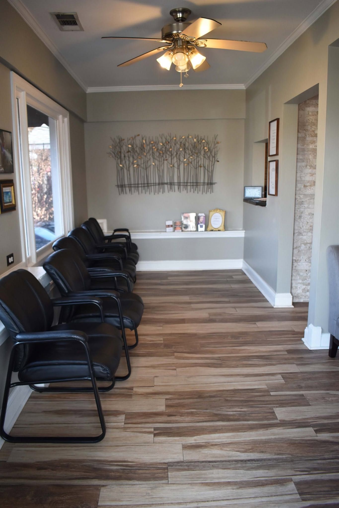 Waiting room with black chairs, wood-look floor, ceiling fan, and wall art in a gray hallway.