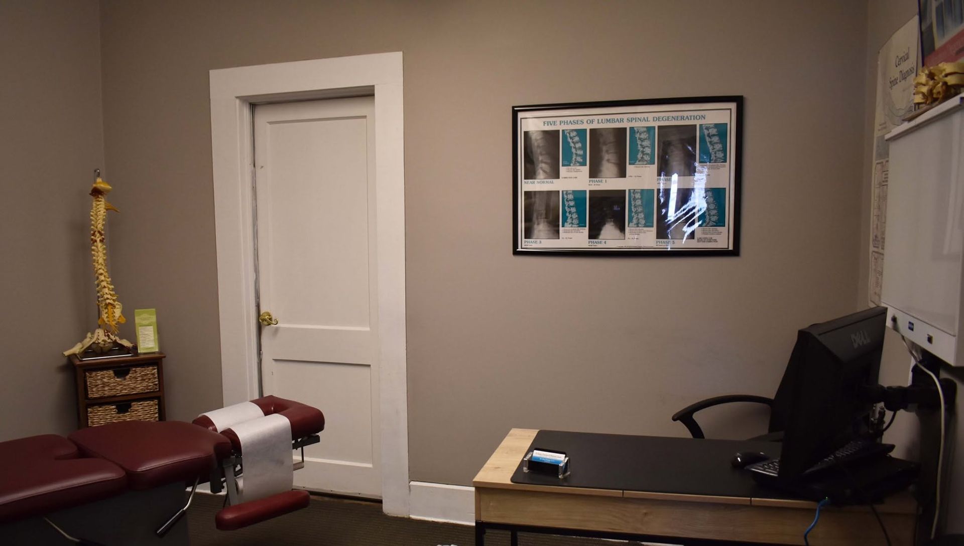 Small office with a desk, computer, maroon exam table, white door, and framed wall art.
