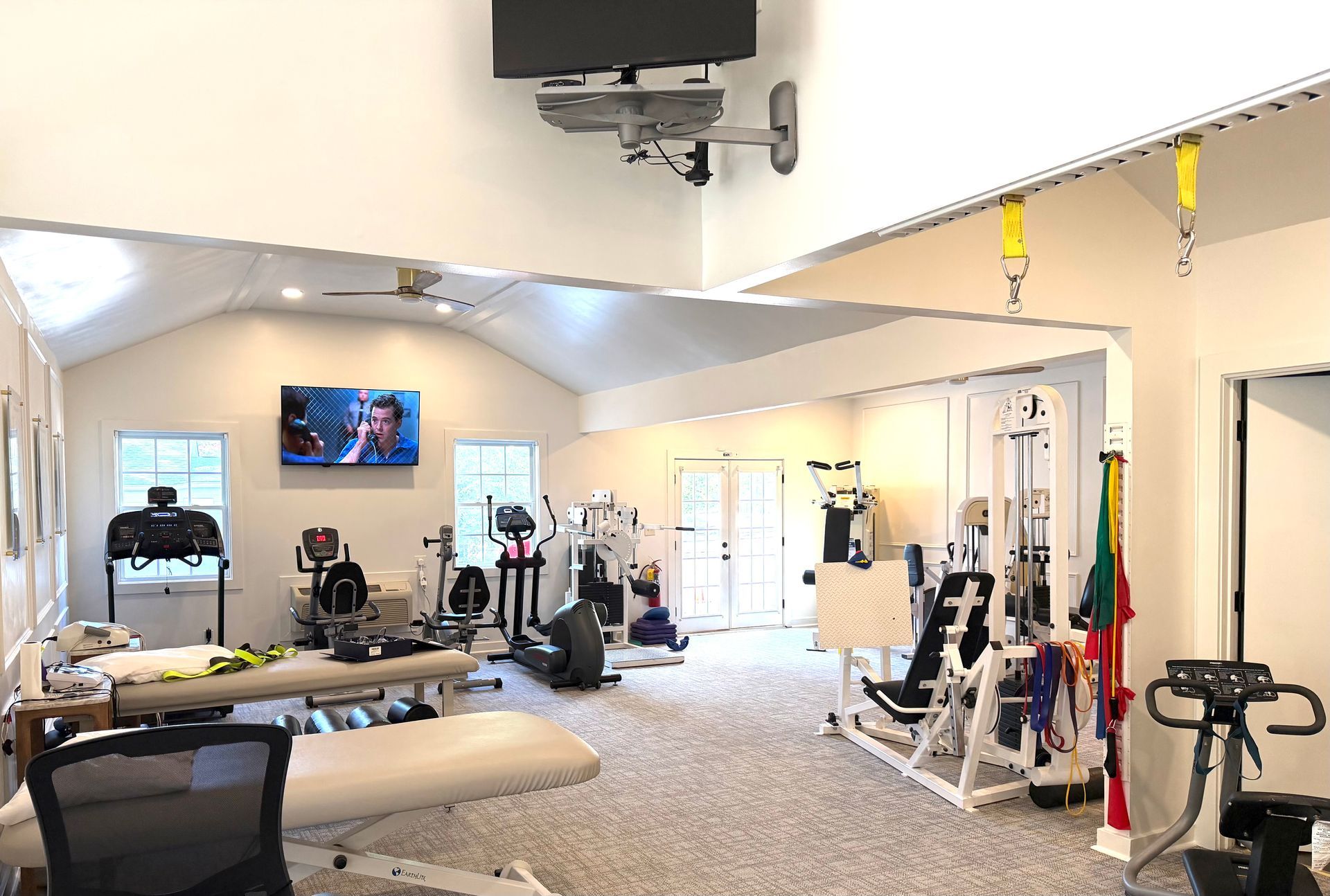 Wide therapy room with exercise equipment, wall-mounted TV, and bright white walls, carpeted floor, and mirrors.