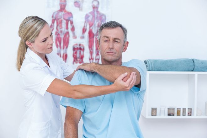 Physical therapist guiding a man’s arm stretch in a clinic, with anatomy charts in the background