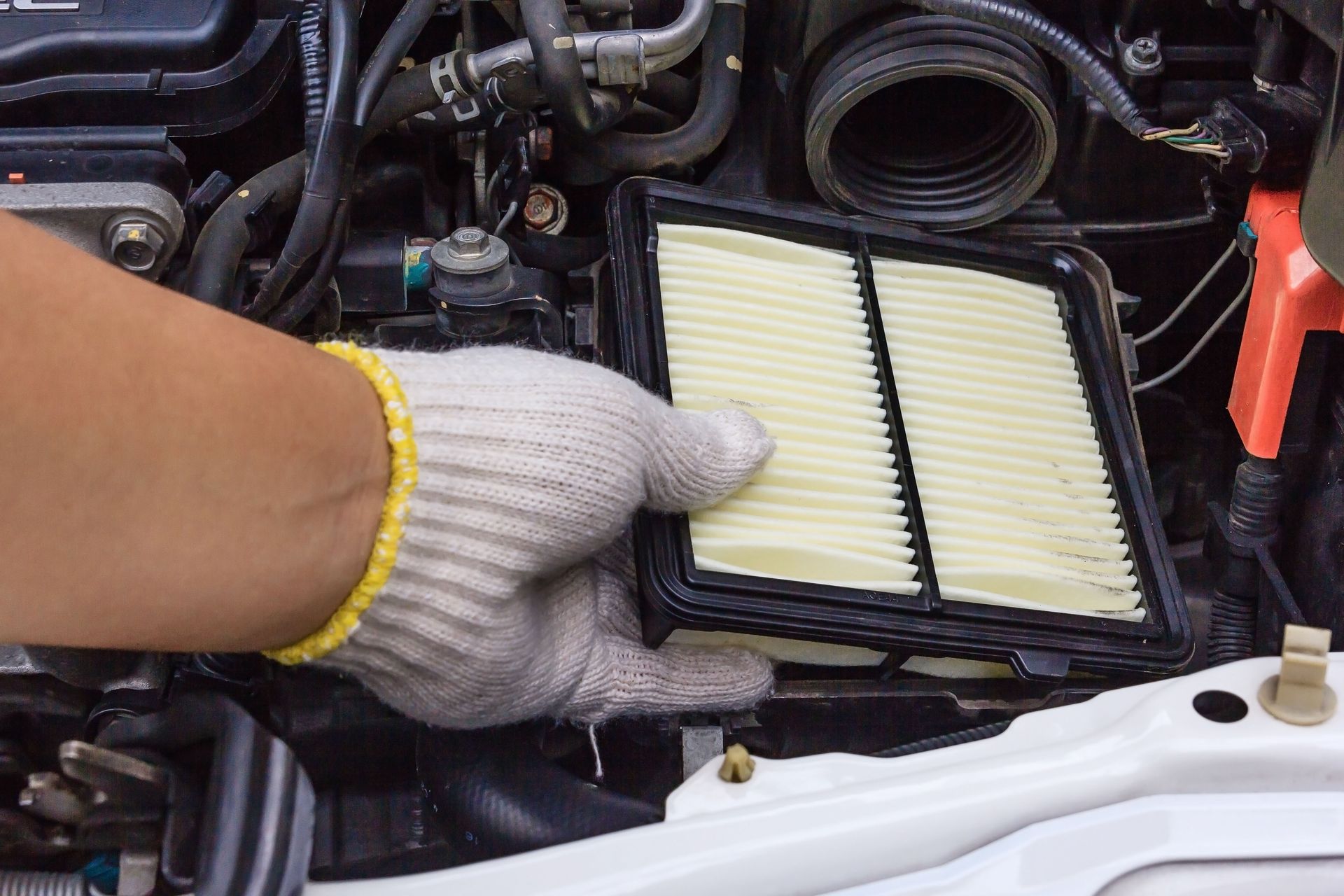 Gloved hand removing an air filter from a car engine. The filter is beige and rectangular.