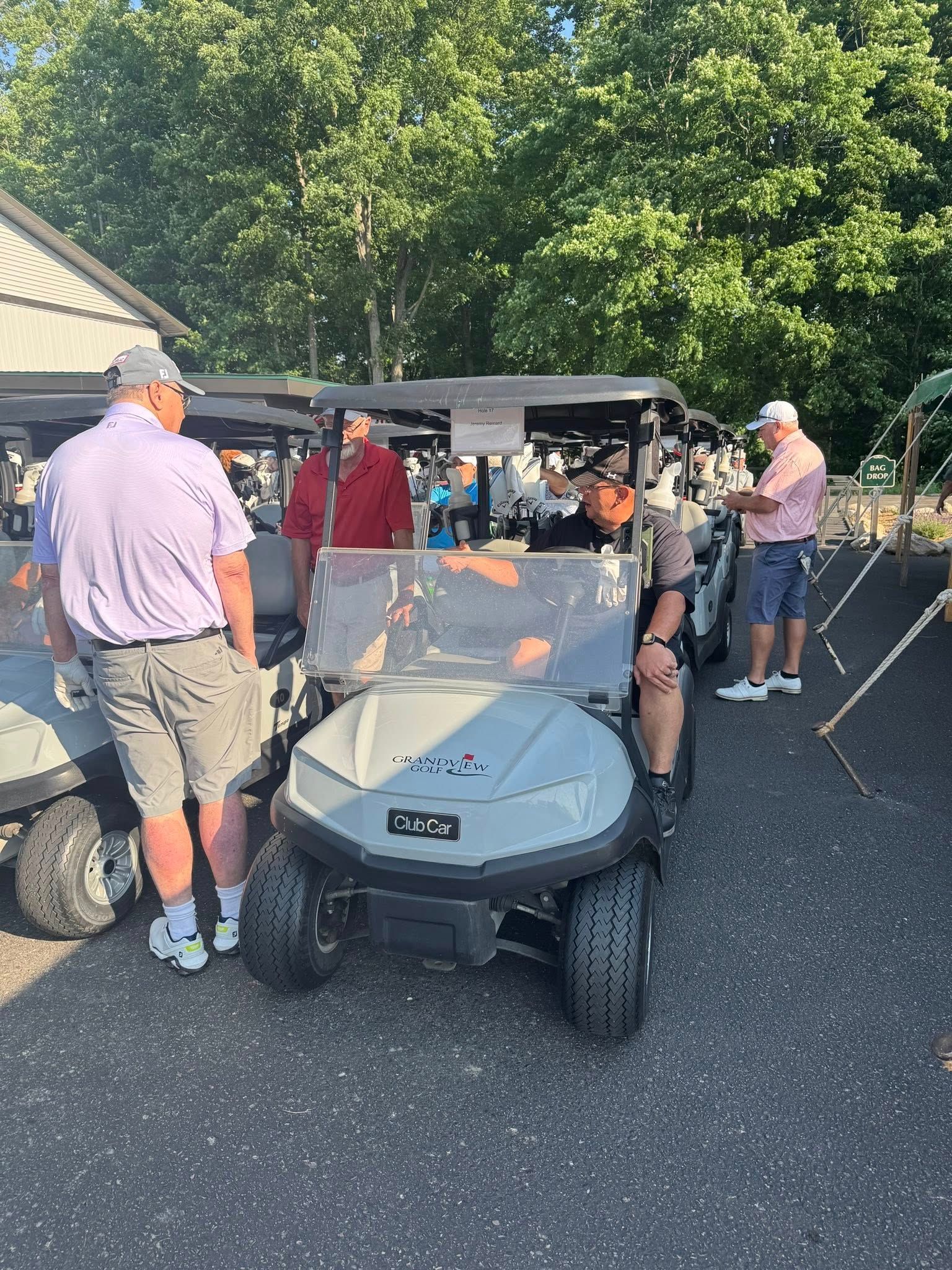 Golfers standing near golf carts on a paved area, trees in the background.