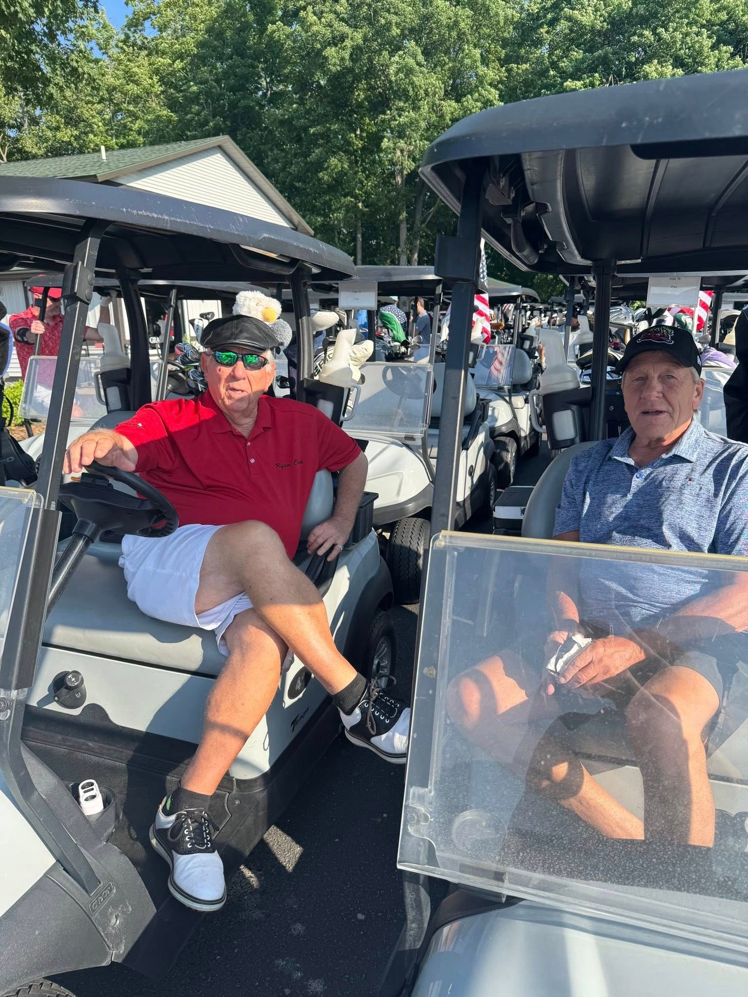 Two men sitting in golf carts on a sunny day. One wears red shirt, white shorts, and sunglasses. The other wears a blue shirt and black hat.