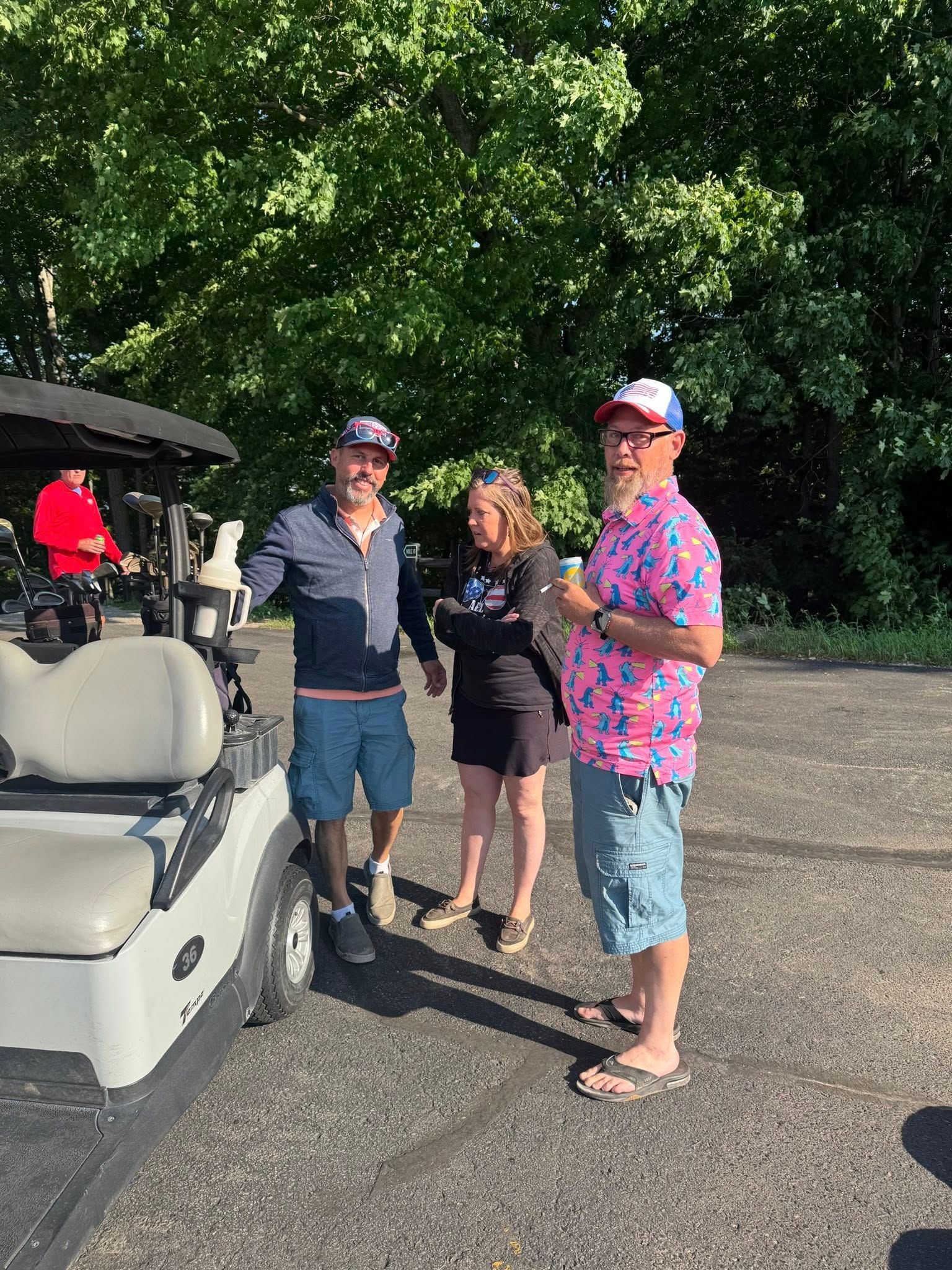 Three people by a golf cart outdoors. Man in pink shirt gestures. Others watch.