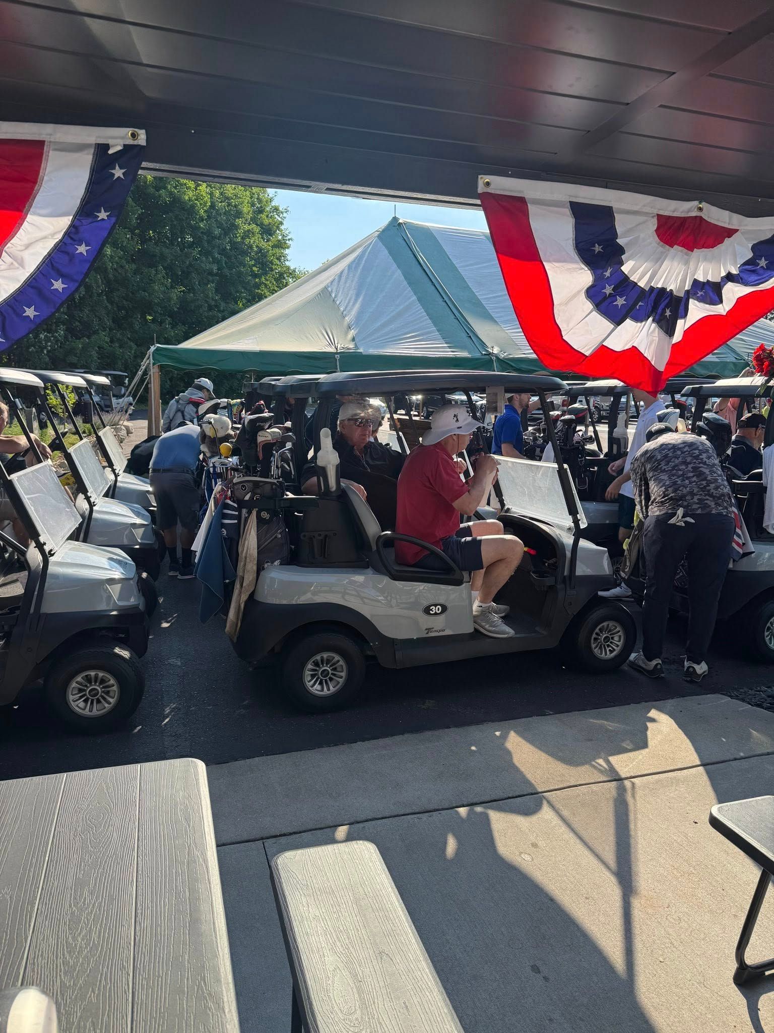 Golfers in golf carts at an outdoor event with a tent and patriotic bunting.