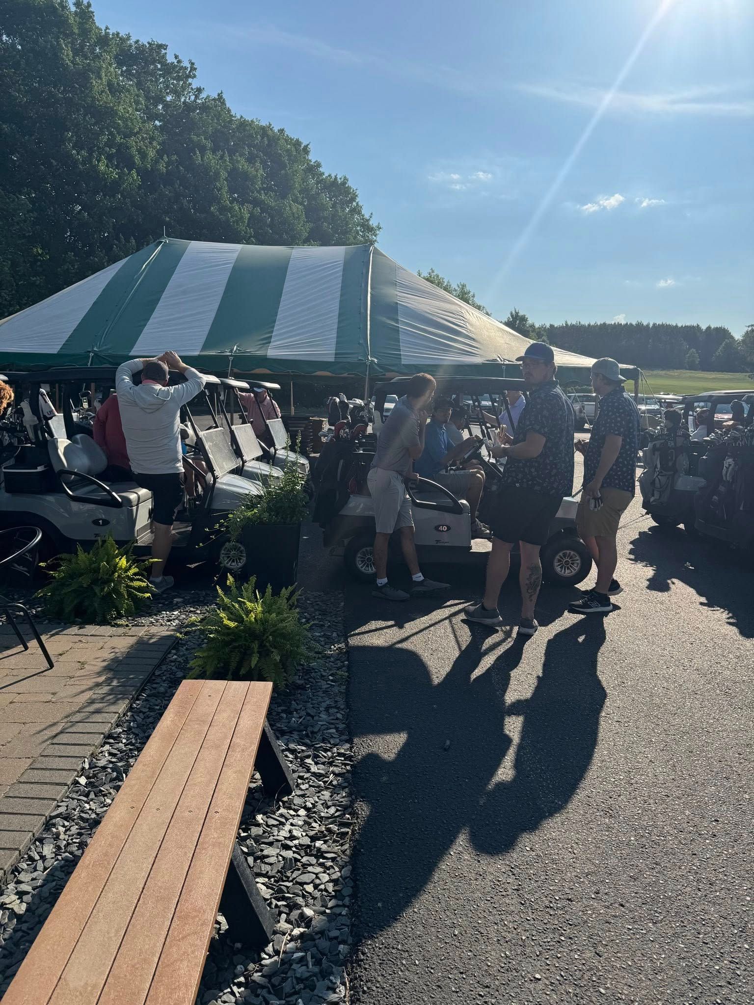 People gather by golf carts under a white and green striped tent on a sunny day.