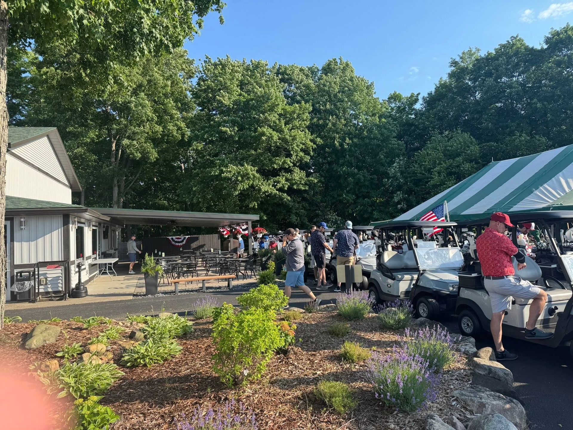 Golfers near golf carts at a clubhouse, under a striped tent. Sunny day, greenery.