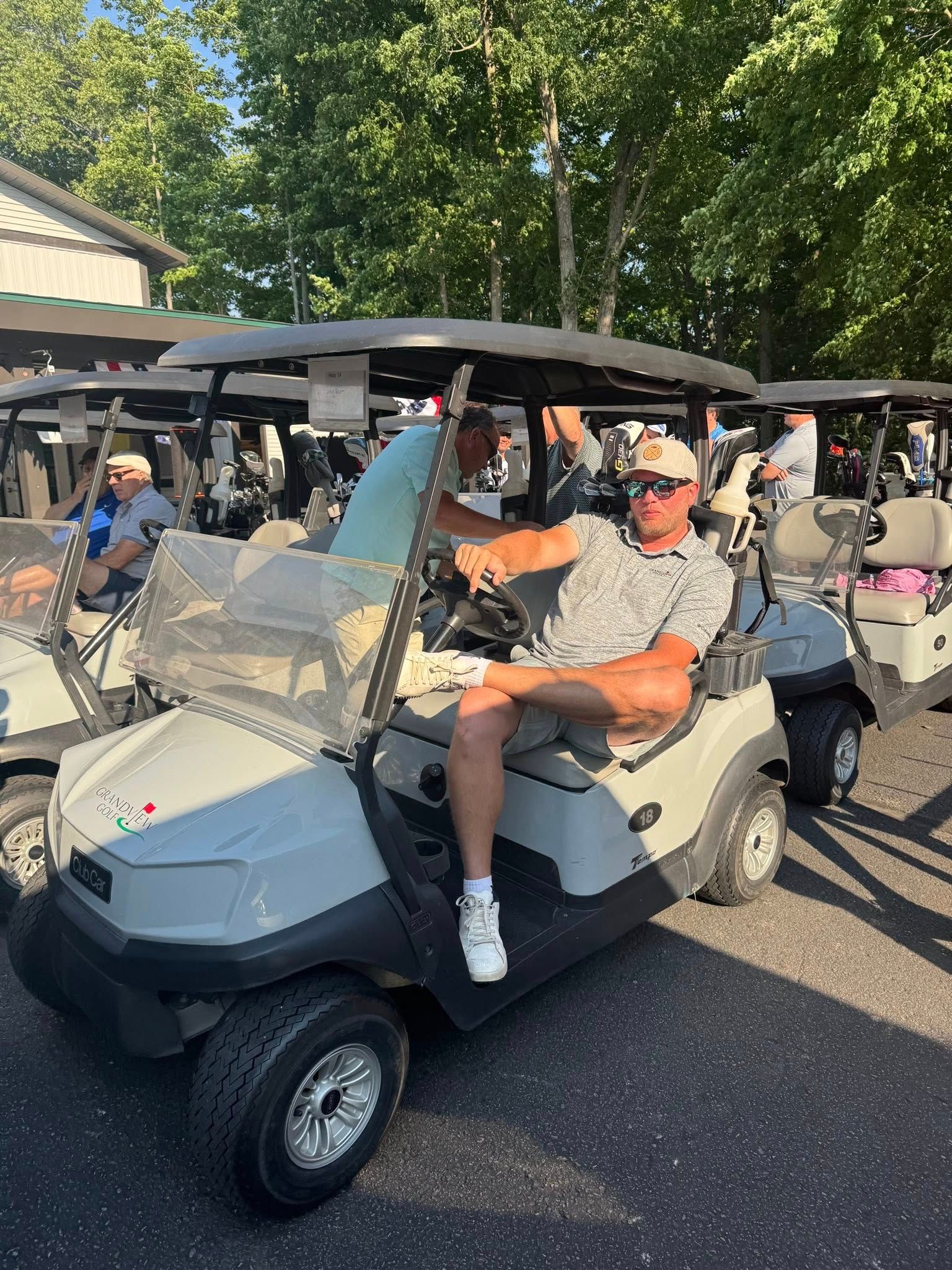 Man sitting in golf cart, smiling. Other carts and trees in background. Outdoors.