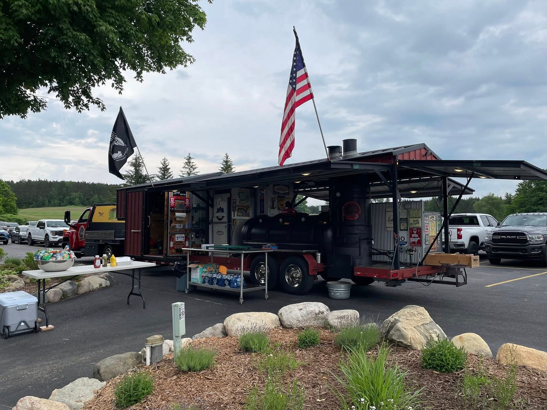 Food trailer with American flag and pirate flag, parked outdoors. Red truck and cars visible.