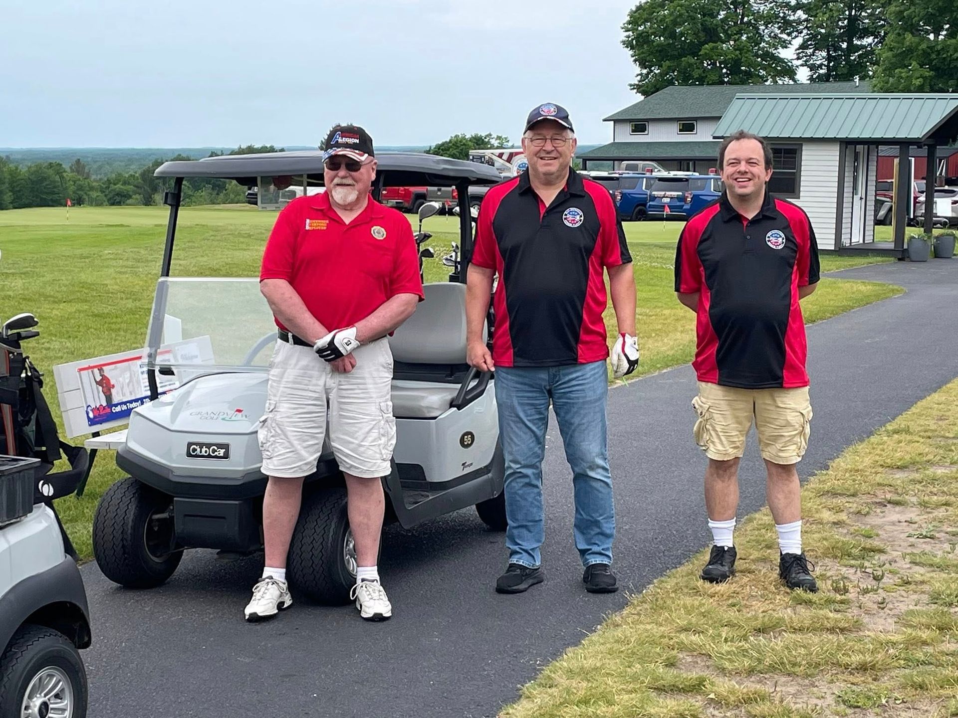 Three men stand by golf carts on a path. They wear red and black shirts, smiling, outdoors.