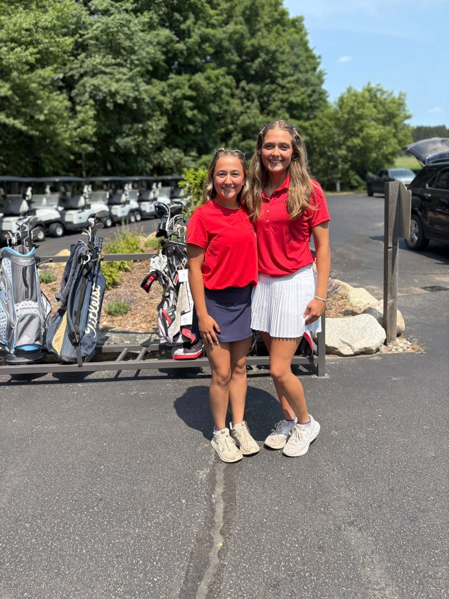 Two young women in red shirts and golf attire, standing near golf carts and bags.