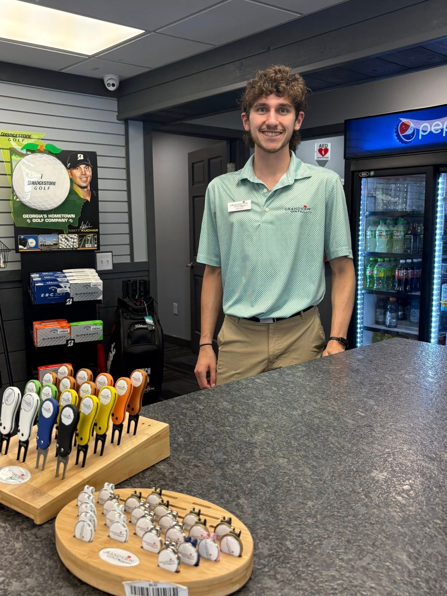 Man in a light green shirt stands behind a counter in a store, smiling. Golf accessories displayed.