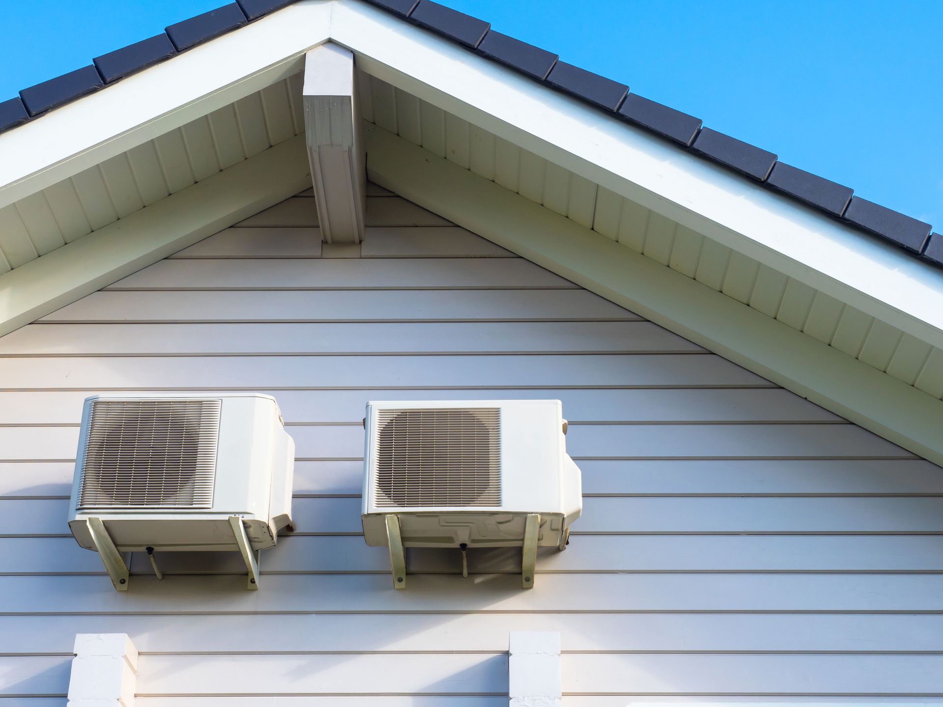 Two white wall-mounted air conditioners on a light gray house exterior under a gabled roof.