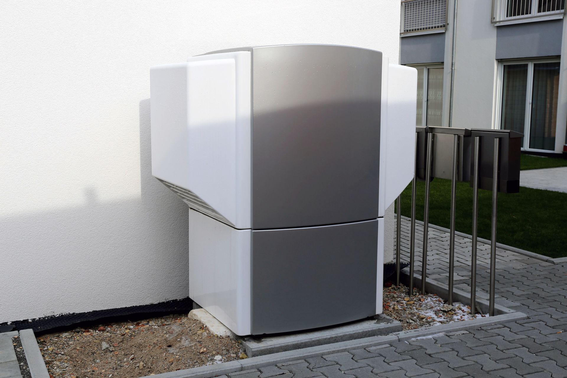 White outdoor equipment unit beside a building, with a small fence and paved walkway nearby