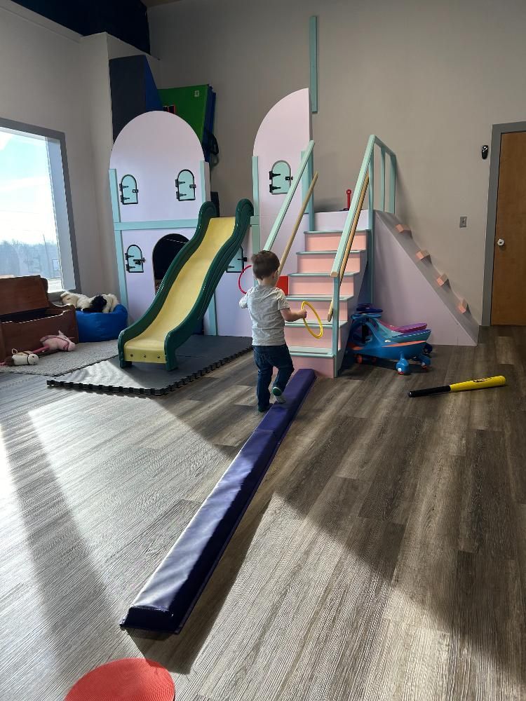 A little boy is standing on a balance beam in a room with a slide and stairs.