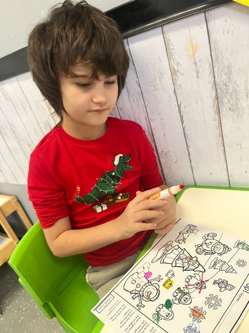 A young boy in a red shirt is sitting at a table holding a marker.