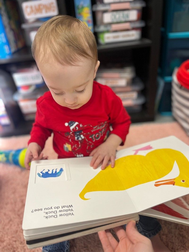 A baby is reading a book with a yellow duck on it.