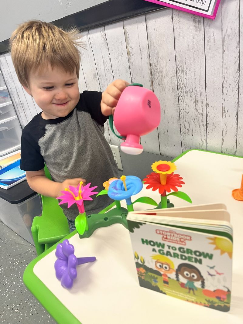 A young boy is watering flowers at a table with a book about how to grow a garden.