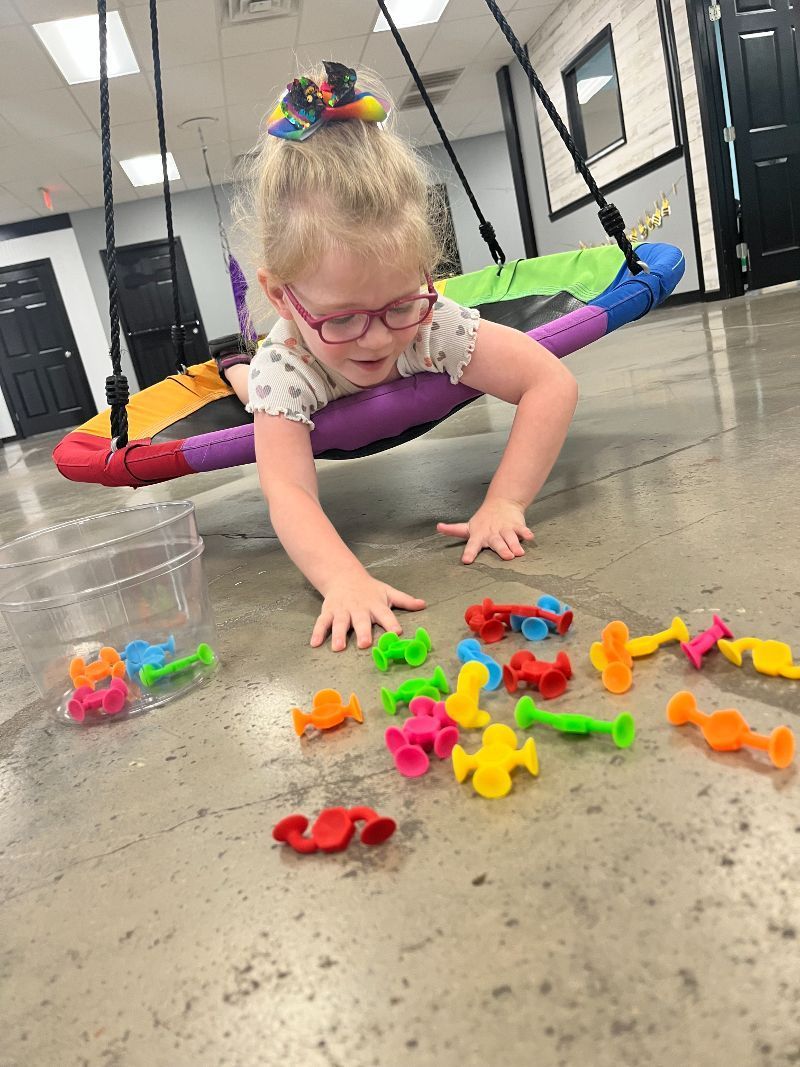A little girl is playing with toys on the floor while sitting on a swing.