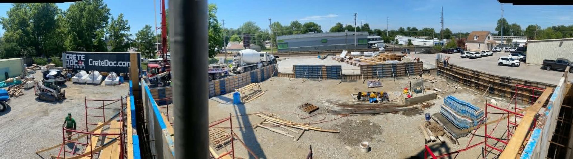 Panoramic construction site with workers, heavy equipment, dirt piles, and a distant skyline under blue sky.