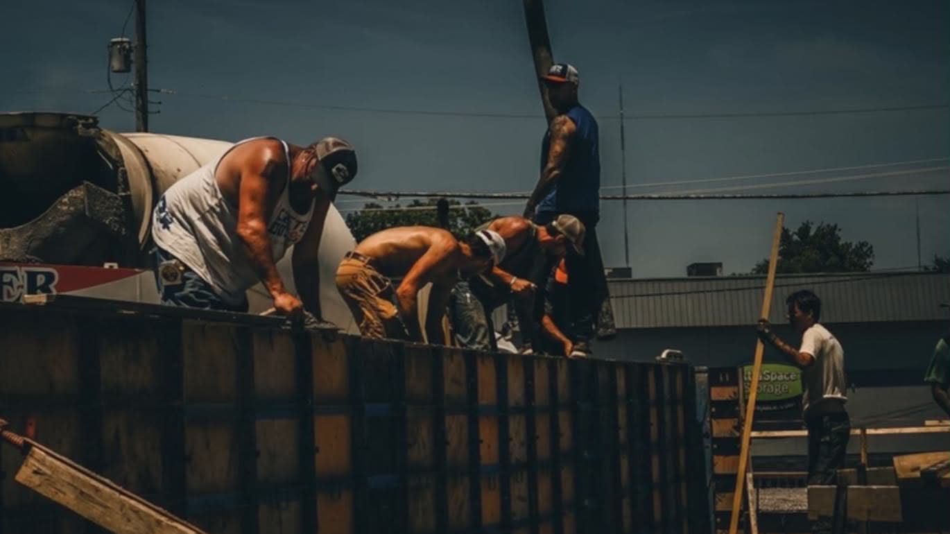 Workers on a rooftop under construction, crouching and bending over materials beside a ladder and fence.