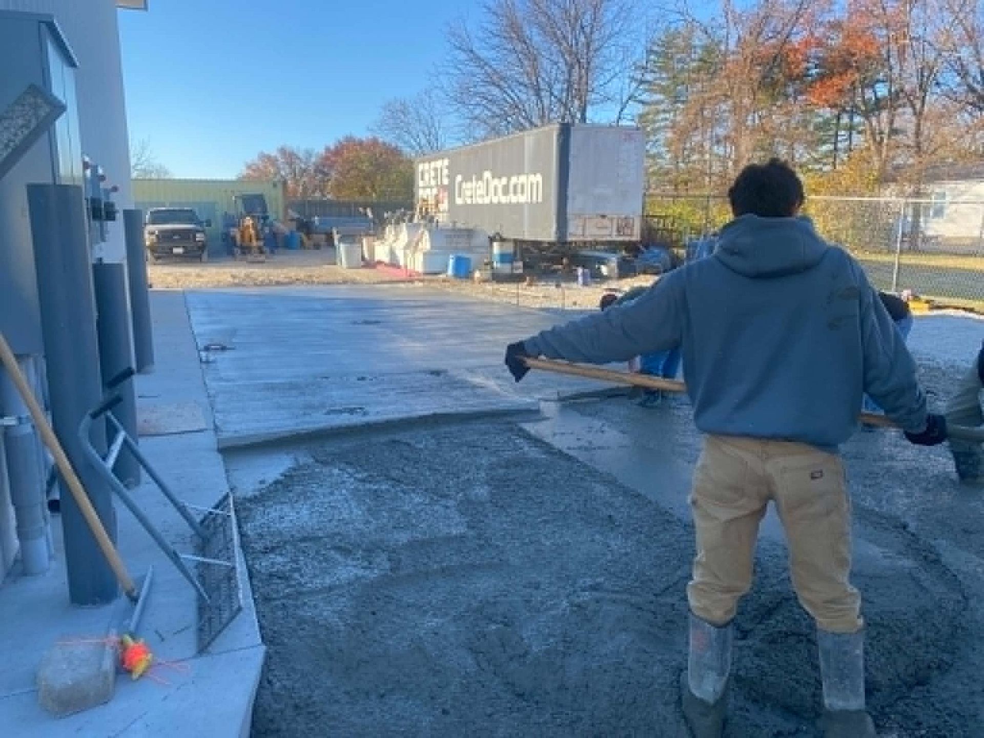 Person in a blue hoodie standing in a parking lot with arms outstretched near a building and trailer.