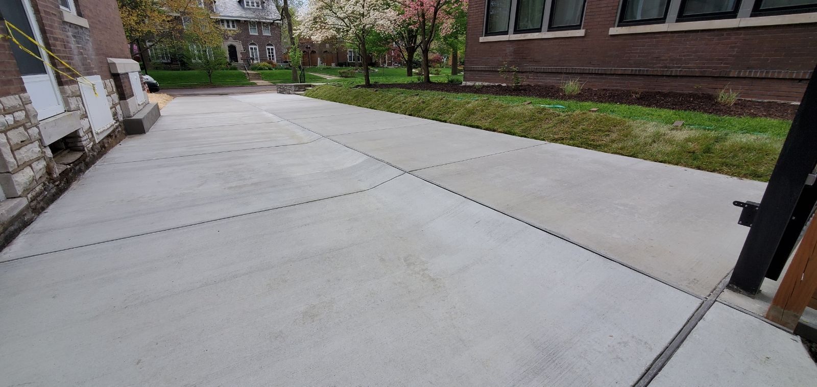 Concrete walkway beside brick building with grass and blooming trees in the background.