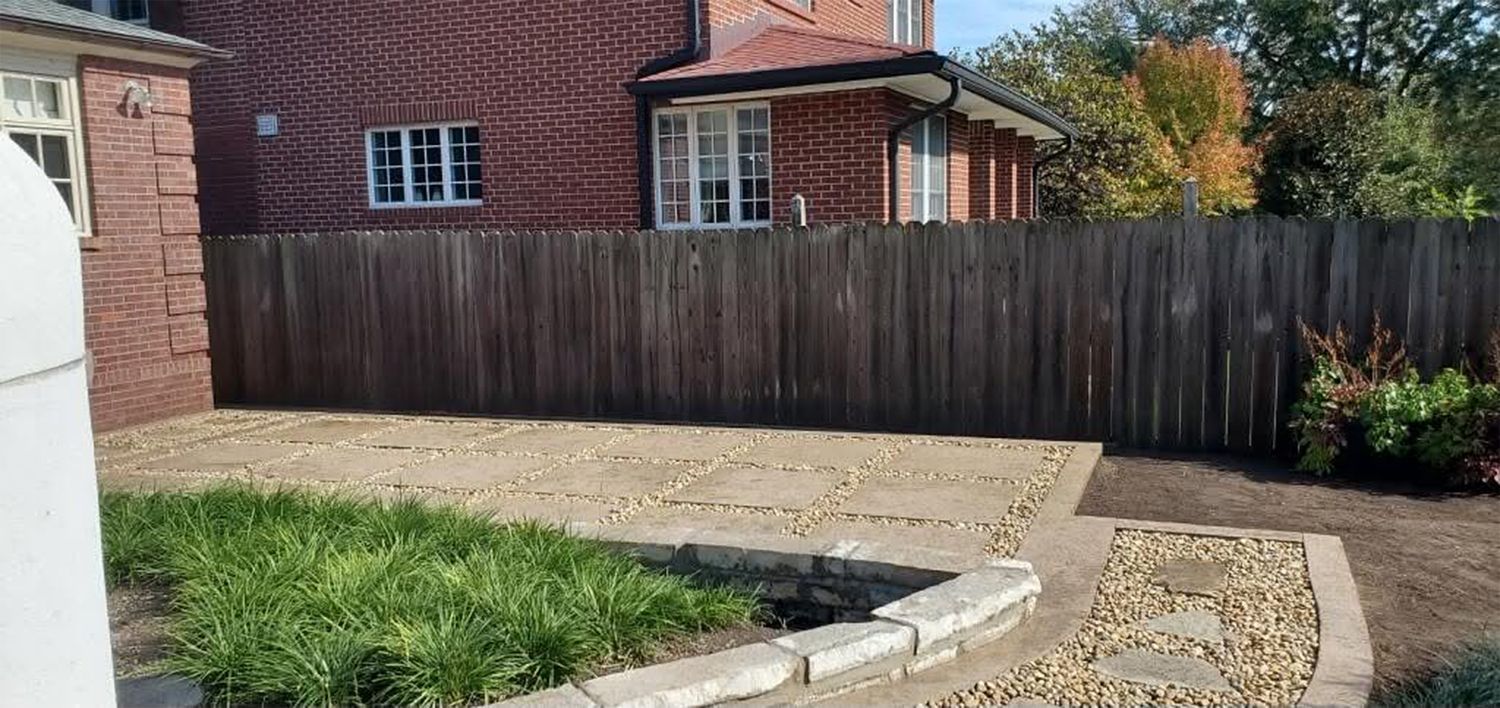 Backyard with curved concrete path, gravel drive, wooden fence, and brick house.