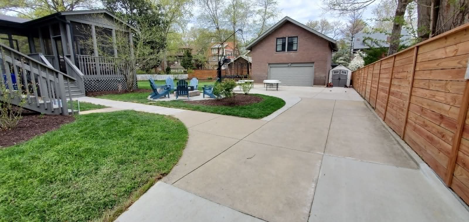 Concrete driveway beside a wooden fence, leading to a house and patio with blue chairs in a landscaped yard.
