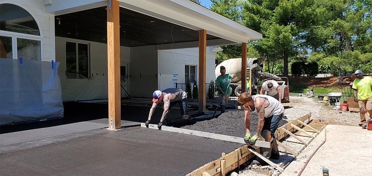 Workers pouring concrete beside a modern building with a covered patio and a sidewalk under construction.