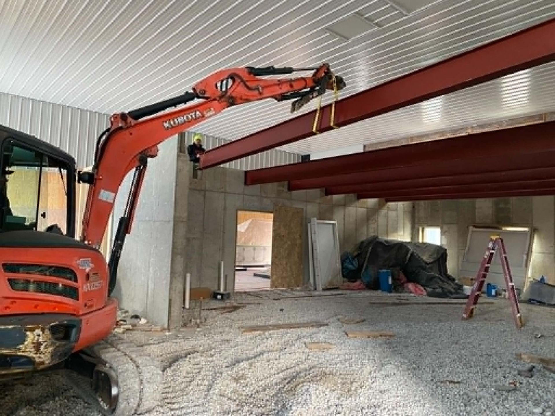 Orange excavator lifting a steel beam inside an unfinished building with concrete walls and debris.