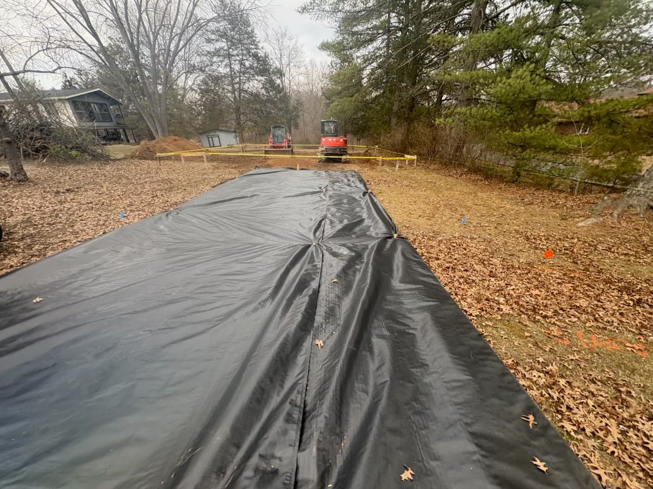 Black tarp laid over a leaf-covered yard with trees and a tractor in the distance.
