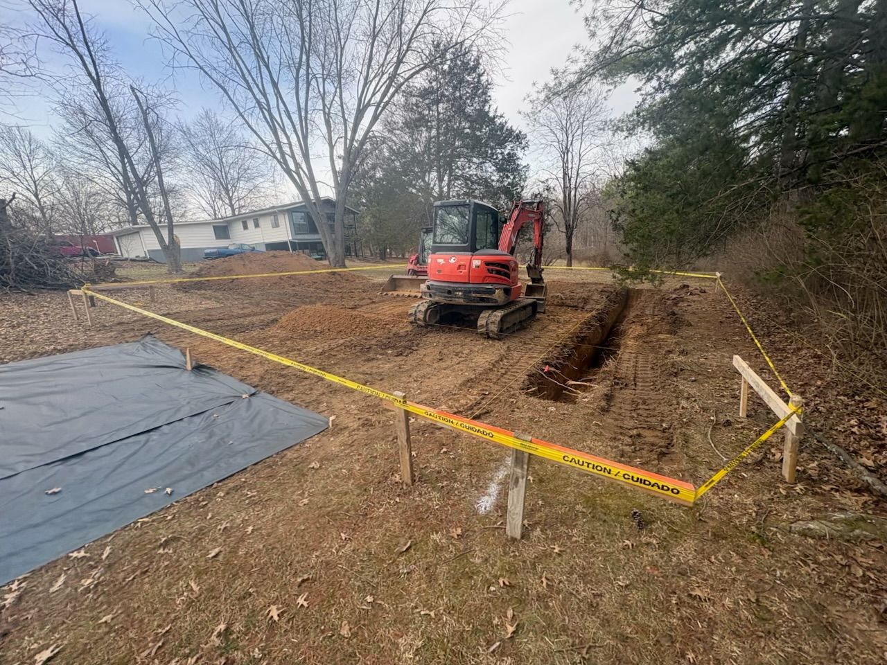Small red excavator digging a trench in a taped-off yard beside a black tarp under bare trees.