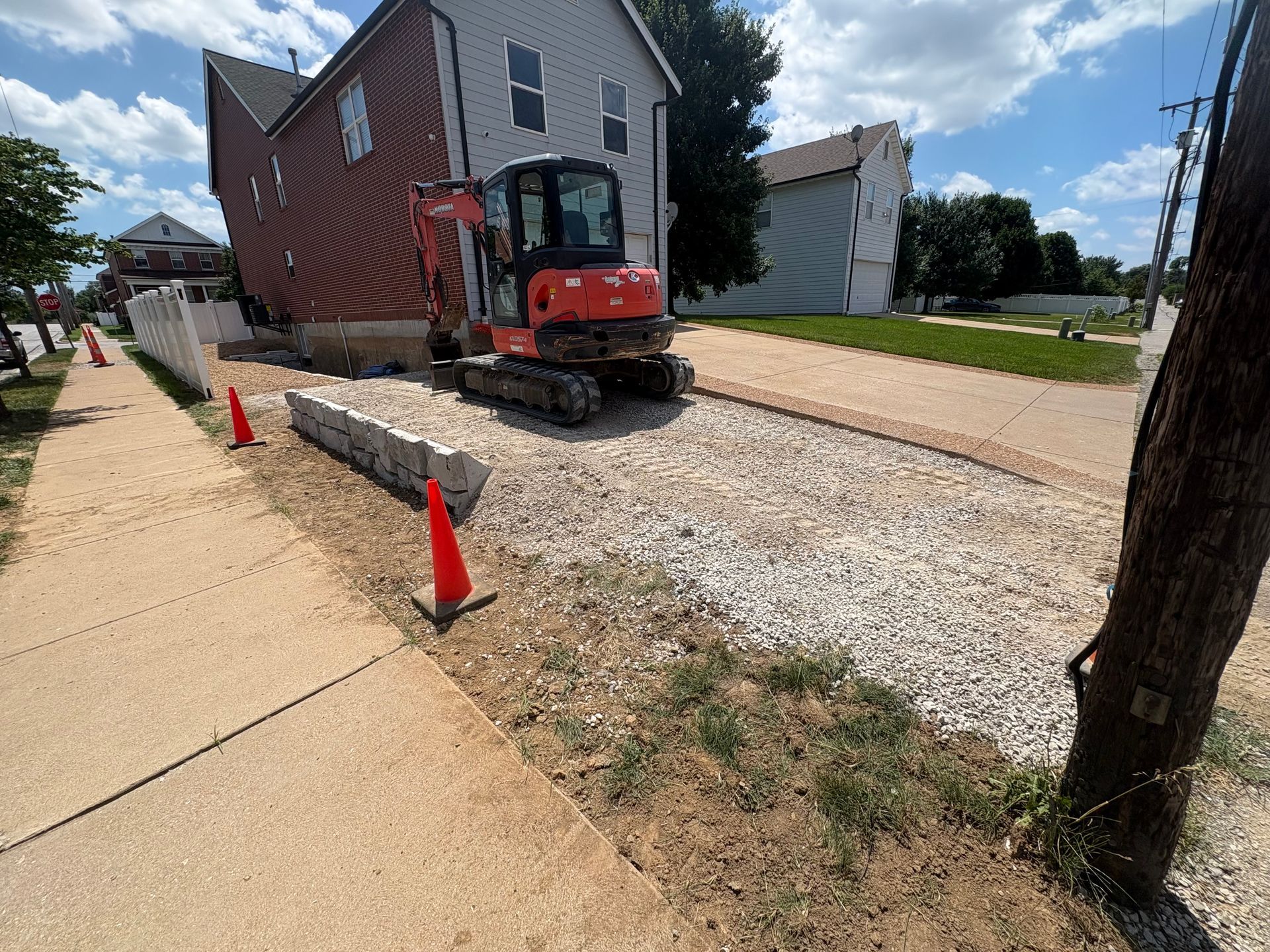 Red excavator working on a gravel driveway beside a house, with orange cones along the sidewalk.