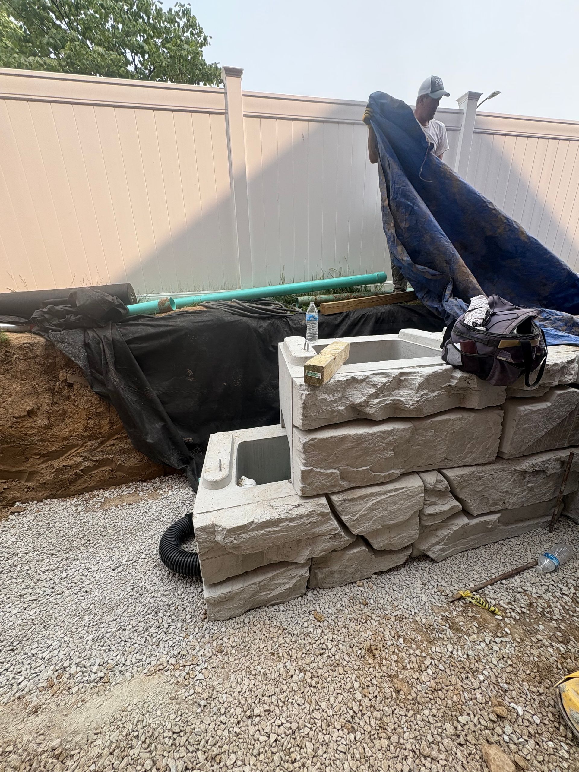 Outdoor yard with a stacked stone retaining wall and black tarp draped over debris beside a fence.