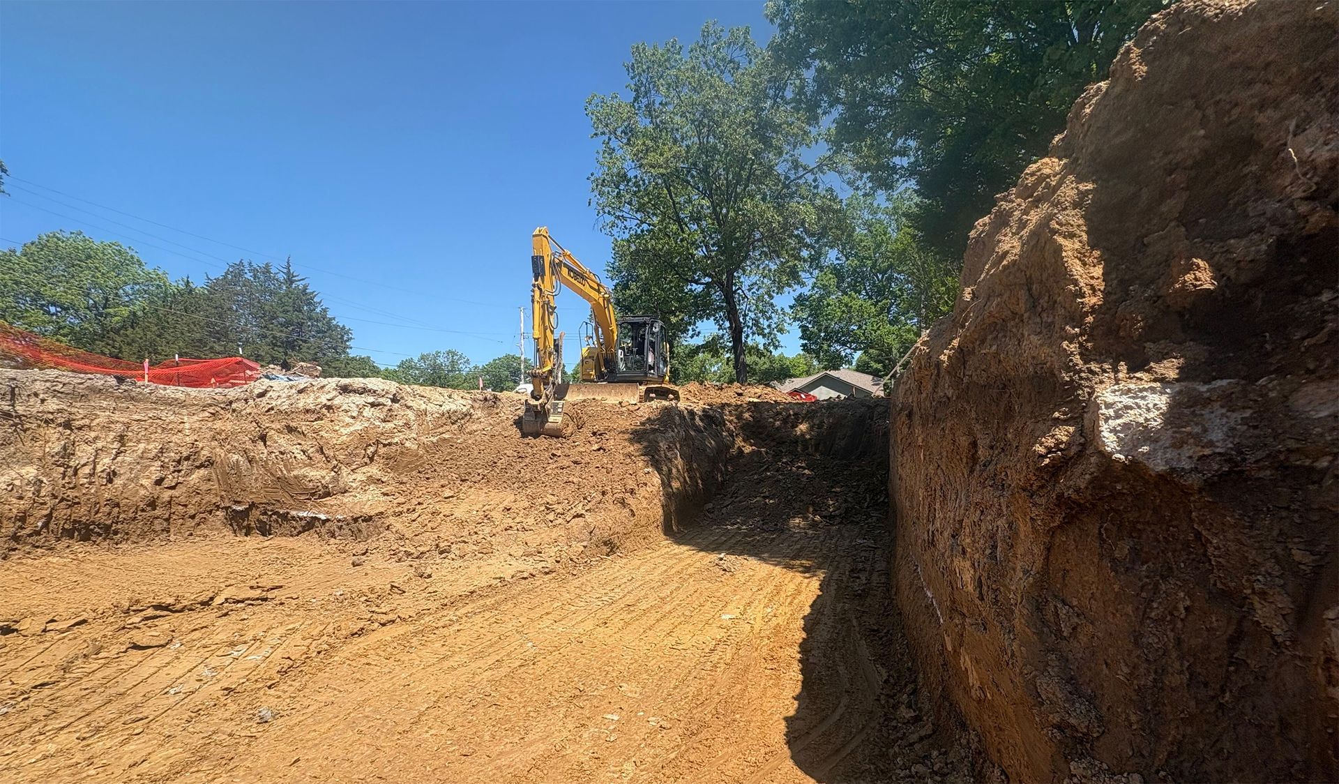 Excavator digging a dirt trench at a construction site under a clear blue sky.