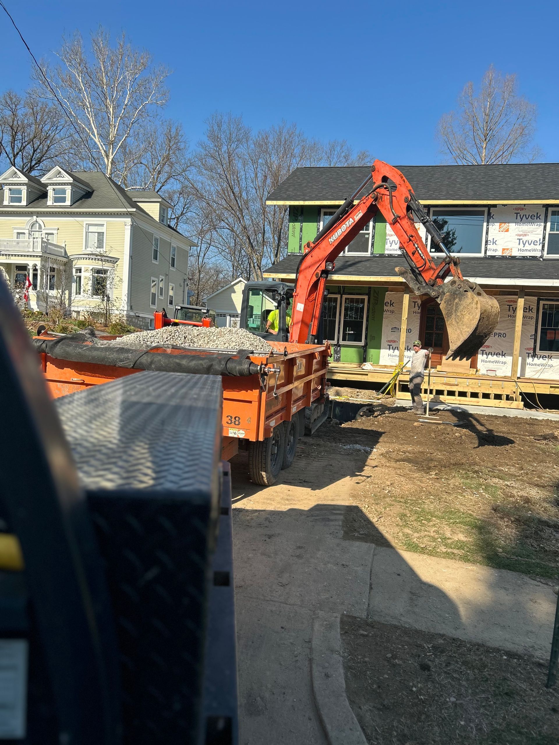 Excavator loading debris into a dumpster in a residential front yard beside a house under renovation.