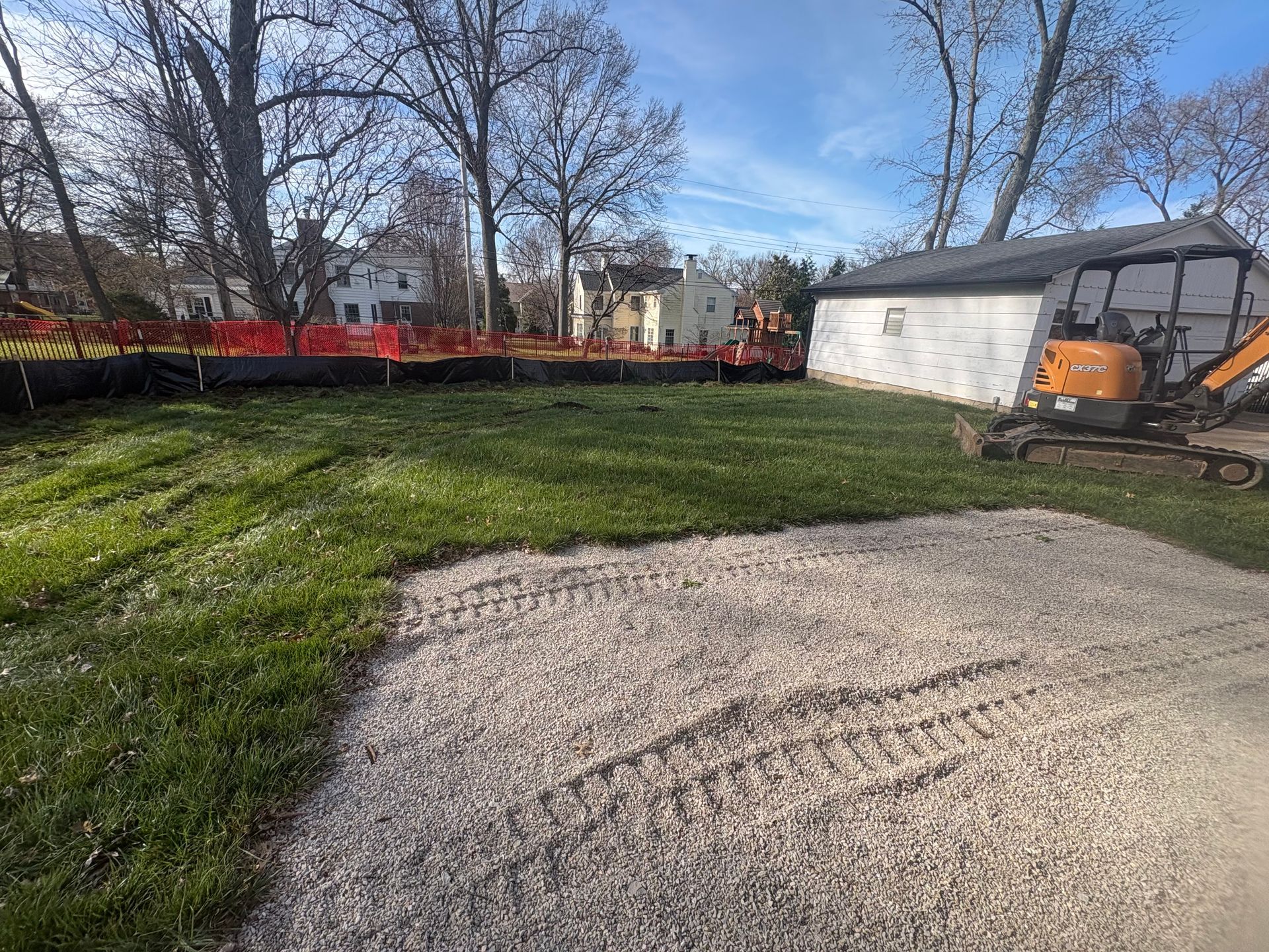 Gravel driveway and grassy yard beside a white garage, with bare trees and a blue sky.