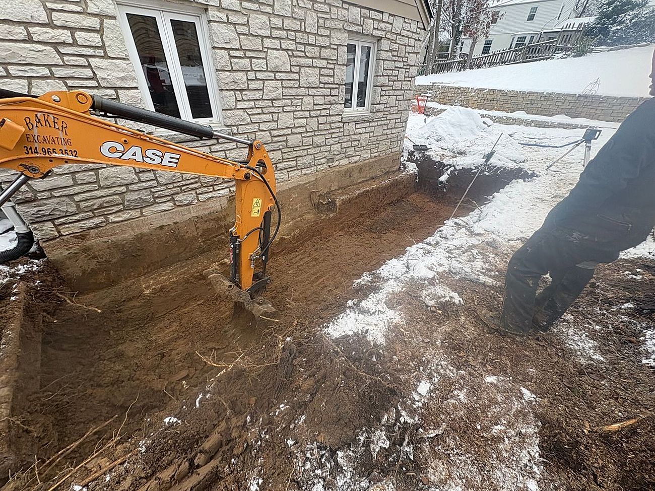 Orange excavator digging beside a stone house in snowy yard, with a person standing nearby.