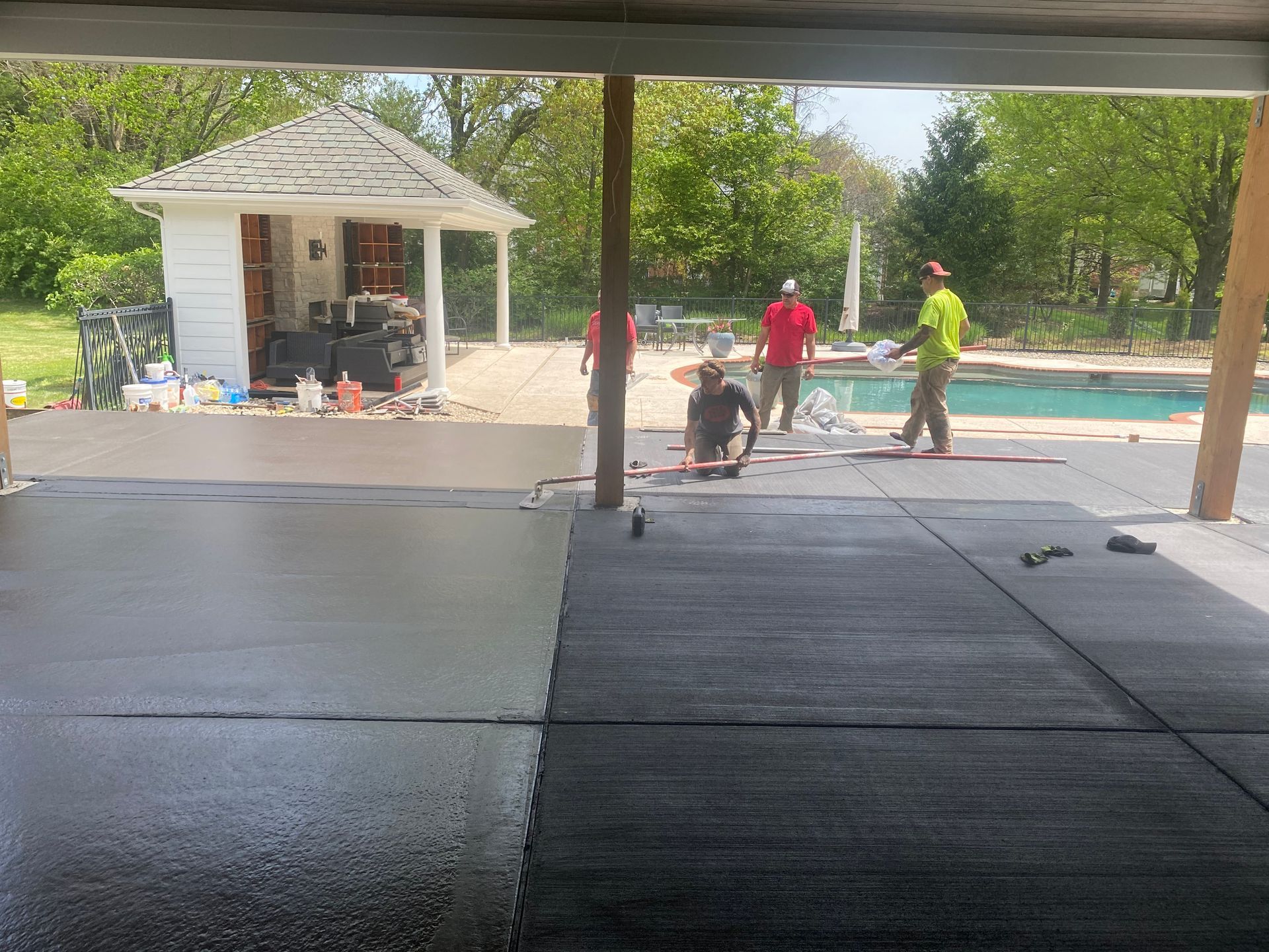Workers resurfacing a backyard patio beside a swimming pool and small shed.